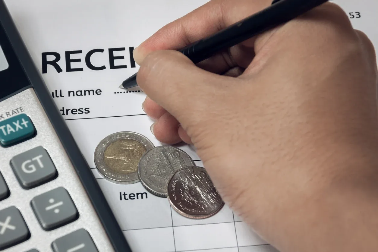 Mão segurando uma caneta sobre moedas e um celular com texto parcial “RECE…”, simulando planejamento financeiro e conciliação de recebíveis para controlar pagamentos e recebimentos