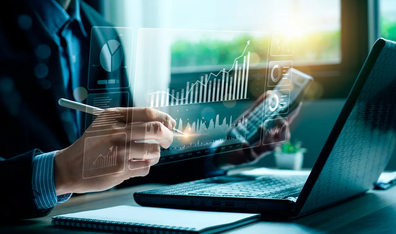 Profissional usando tablet com gráficos e números financeiros sobrepostos, simbolizando plano de contas e análise contábil em escritório com computador e documentos na mesa.